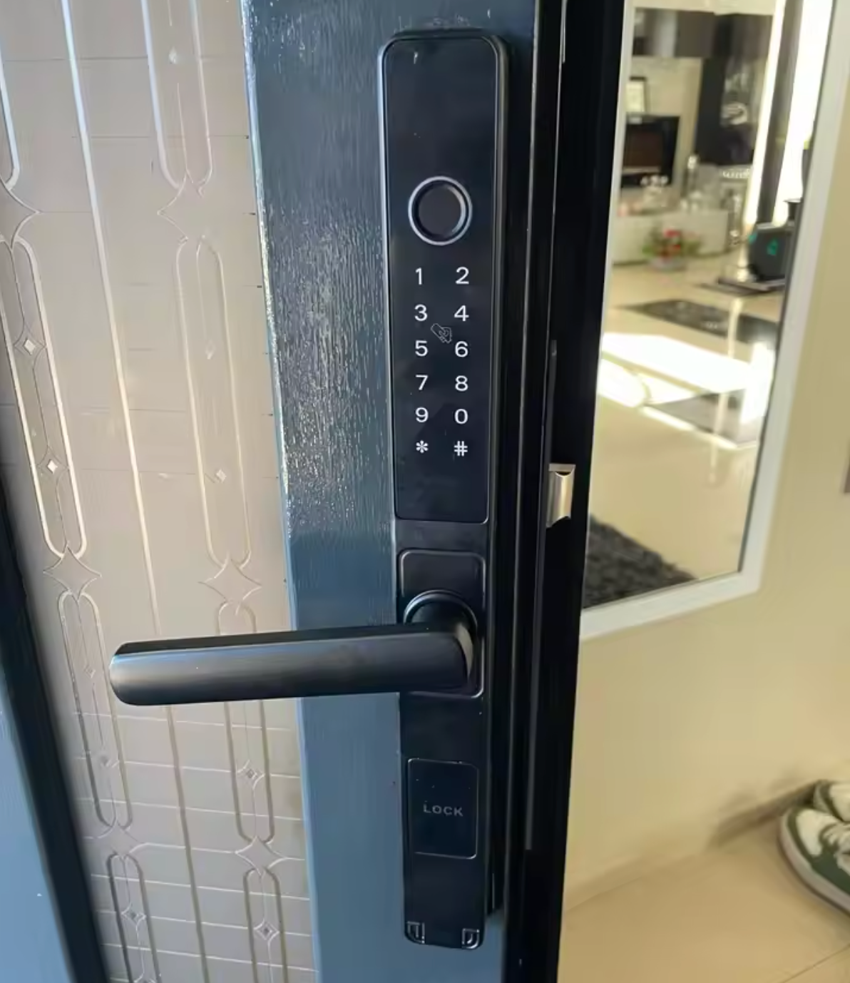Black smart door lock with keypad and lever handle fitted to a glazed entry door, hallway behind
