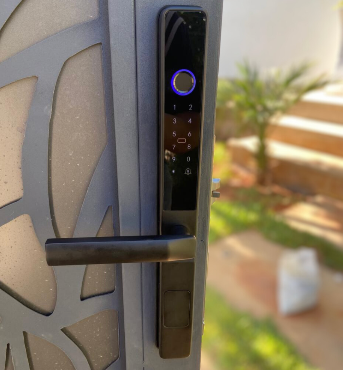 Black smart door lock with a lit fingerprint ring, fitted to a carved timber door with a garden behind